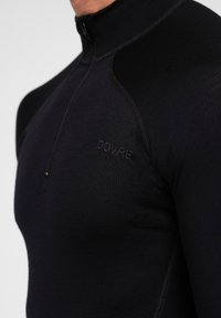 Schwarzes Langarm-Zip-Up-Shirt aus einem strukturierten Stoff, mit kontrastierenden Schulterpaneelen und einem gestickten Logo auf der Brust.