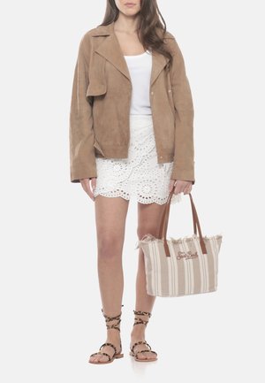 Donna che indossa una giacca in camoscio marrone, gonna in pizzo bianca, top bianco, sandali con stampa leopardata e che tiene in mano una borsa tote beige a righe.