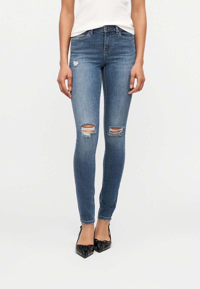 Γυναίκα που φοράει μπλε σκισμένο στενό τζιν, λευκή μπλούζα και μαύρα παπούτσια με μυτερή μύτη, στέκεται μπροστά σε ένα απλό φωτεινό φόντο.