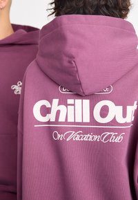 Lilla hettegensare laget av bomull, med stor hvit grafisk tekst "Chill Out" og mindre tekst "On Vacation Club" på baksiden.