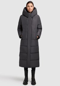 Donna in piedi che indossa un lungo cappotto imbottito nero con colletto alto e stivali neri, su uno sfondo grigio chiaro uniforme.