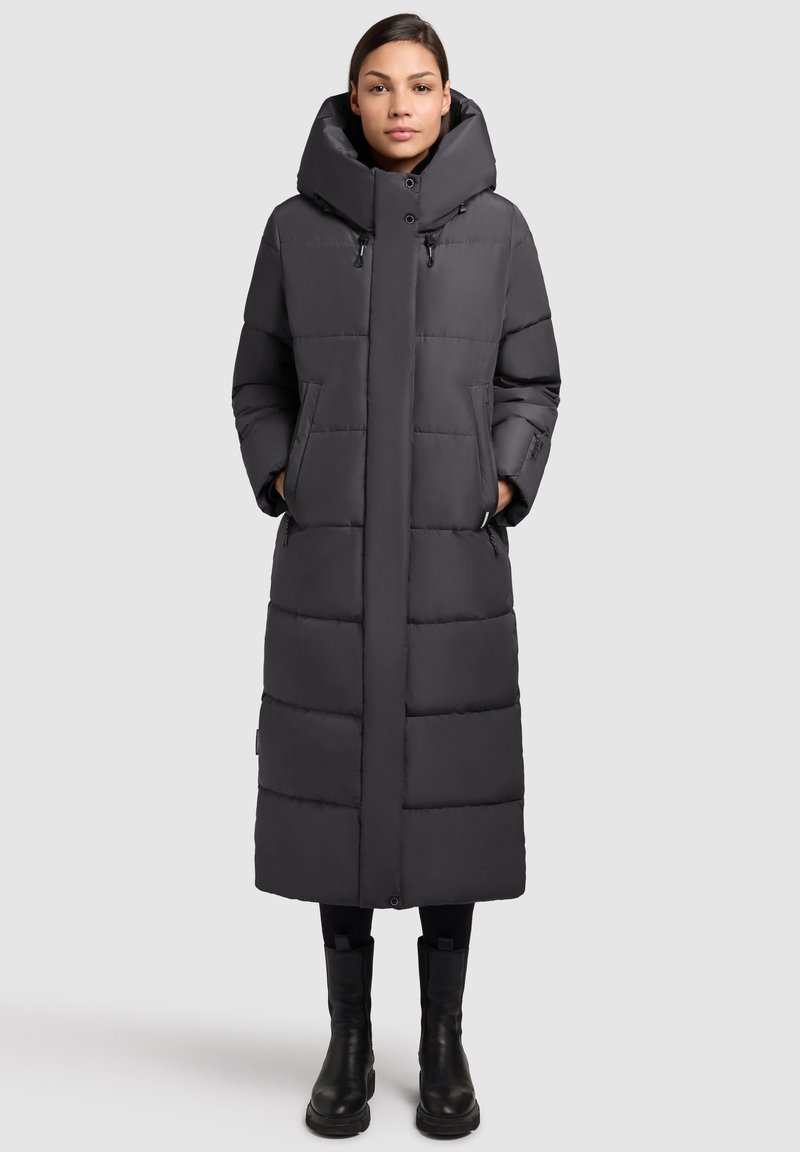 Donna in piedi che indossa un lungo cappotto imbottito nero con colletto alto e stivali neri, su uno sfondo grigio chiaro uniforme.