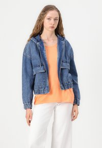 Denim jakna do pola s kapuljačom, elastičnim manžetama i porubom, svijetloplave boje, s dva prednja džepa. Nosi se preko narančaste majice.