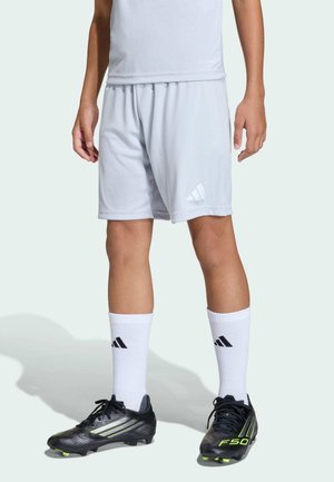 Osoba ubrana w jasnoszare sportowe spodenki, białe skarpety Adidas i czarne korki piłkarskie Adidas z żółtymi akcentami, stojąca na jasnym tle.