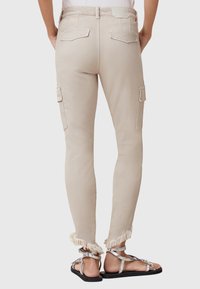 Pantalones cargo beige con un ajuste slim, dobladillo deshilachado, dos bolsillos laterales y dos bolsillos traseros, combinados con sandalias planas plateadas.