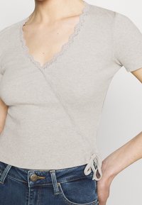 Světlý šedý žebrovaný top s krátkým rukávem a krajkovým výstřihem ve tvaru vlnky, s překládaným designem a detailem uvázání na boku.