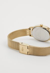 Montre-bracelet en mailles dorées avec un boîtier rond lisse et un bracelet texturé. Le dos présente un logo et un compartiment transparent avec les détails.