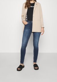 Blazer beige sobre una camiseta gráfica negra, combinado con jeans ajustados de color azul oscuro y sandalias negras tipo slide. Tela suave y diseño a medida.