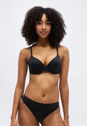 Femeie cu păr creț, purtând un sutien negru și lenjerie asortată, pe un fundal simplu gri deschis, stând în picioare și cu fața spre înainte.