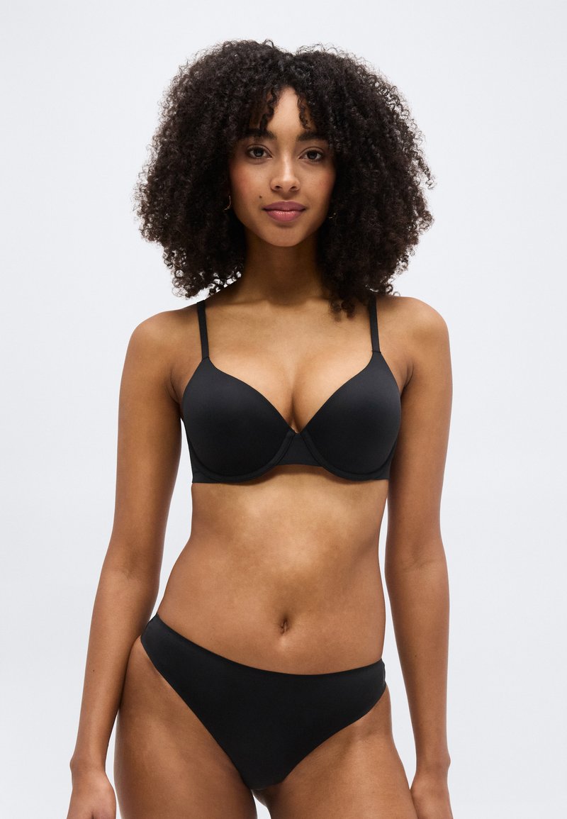 Γυναίκα με σγουρά μαλλιά που φοράει μαύρο σουτιέν και αντίστοιχο εσώρουχο, σε απλό ανοιχτό γκρι φόντο, όρθια και κοιτάζοντας μπροστά.