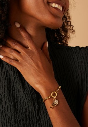 Goldene Kettenarmband mit einem runden Anhänger und einem Haken-Detail, getragen an einem Handgelenk mit strukturiertem Hautbild, vor einem dunklen, gerippten Stoffhintergrund.