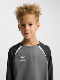 Kind met krullend blond haar dat een grijs en zwart Hummel sportshirt met lange mouwen draagt, voorover gericht op een effen achtergrond.