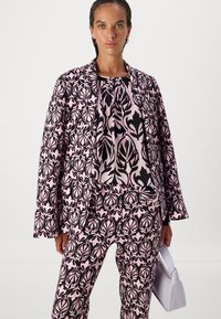 Rosa und schwarzes gemustertes Anzug mit floralen Designs. Enthält eine Jacke mit langen Ärmeln, passende Hose und eine weiße Handtasche. Glatte Textur.