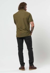 Homme aux cheveux bouclés, debout, de dos, portant un polo vert olive, un jean noir, des chaussures marron et un tatouage sur le bras droit.