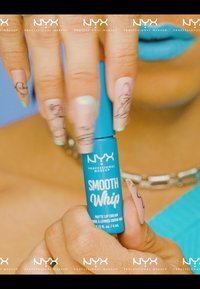 Een blauwe tube van NYX Smooth Whip matte lipcrème, 4 ml, met witte tekst. De hand heeft lange, nude nagels met zwart lijnkunstontwerp.