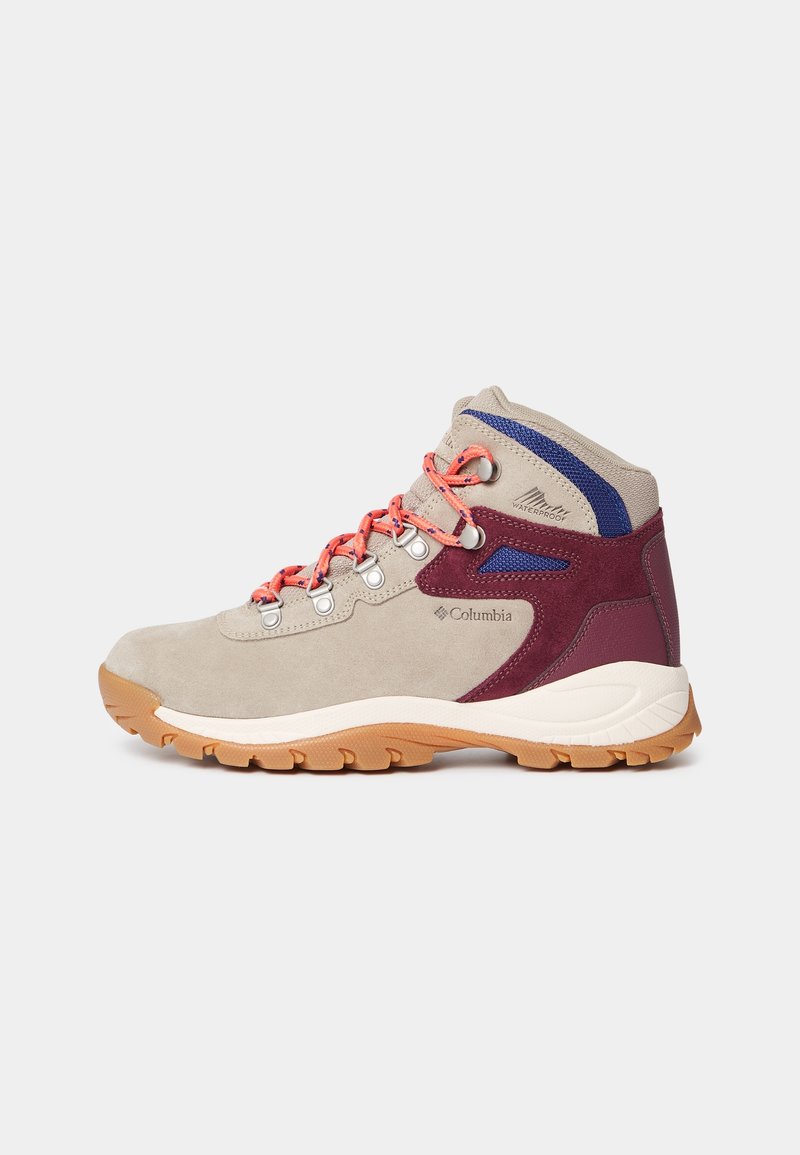 Botte de randonnée beige avec des accents bordeaux et bleus, lacets orange, semelle marron robuste, étanche avec le logo Columbia sur le côté.