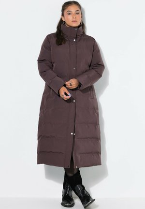 Mujer de pie con un abrigo largo acolchado marrón oscuro con cuello alto, botas negras y anillos en los dedos sobre un fondo liso y claro.