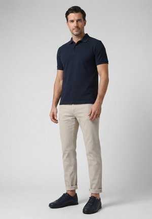 Muž v tmavě modrém polu, béžových vyhrnutých kalhotách a černých teniskách stojící před jednobarevným šedým pozadím.