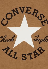 Material maro prezintă o stea albă mare în centru, înconjurată de text negru cu cuvintele „CONVERSE”, „Chuck” și „ALL STAR”.
