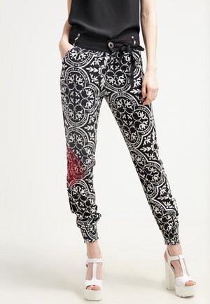 Vrouw draagt zwart-witte patroonbroek met een rood stuk op het linkerbeen, zwarte top en witte plateauhakken, staat met handen in de zakken.