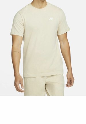 Homme portant un t-shirt beige Nike à manches courtes et un pantalon assorti, debout devant un fond blanc uni.