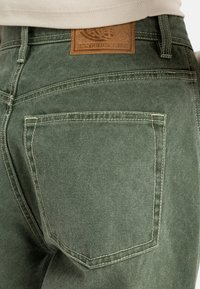Pantalons en denim vert avec une poche arrière, des coutures contrastantes et une étiquette en cuir marron au niveau de la taille.