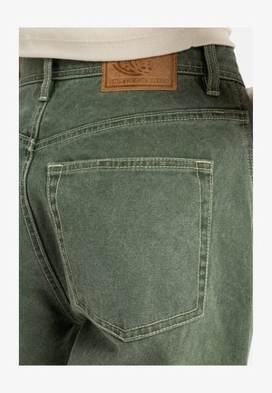 Green denim pants featuring a back pocket, contrasting stitching, and a brown leather brand patch at the waistband.
