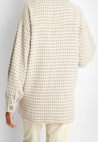 Camisa chaqueta de patrón houndstooth en beige y blanco, con mangas oversized y puños abotonados, que presenta un canesú en la parte posterior y un ajuste relajado.
