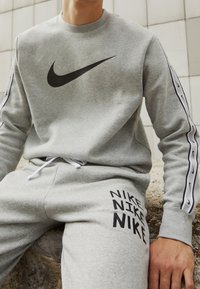 Grå sweatshirt med stort svart Nike-logotyp, med vita accenter på ärmarna. Matchande joggers har svart "NIKE"-textmönster på låret.
