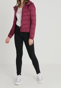 Maroon puffer jacket with a hood, featuring a zip closure and horizontal quilted texture, paired with black skinny jeans and white sneakers.