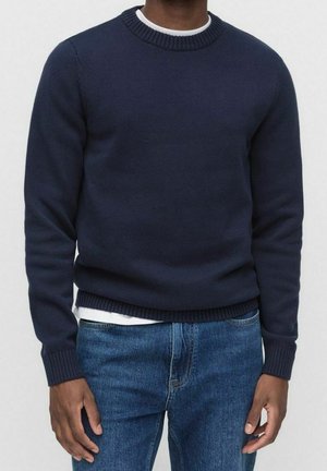 Mann trägt einen dunkelblauen Strickpullover über einem weißen Hemd mit blauen Jeans, steht vor einem einfarbigen hellen Hintergrund.