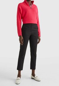 Person med en klar röd zip-up tröja, svarta slim-fit byxor, vita loafers med guldkedjedetalj, som står mot en neutral bakgrund.