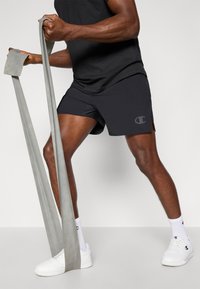 Pantalones cortos deportivos negros con aberturas laterales, banda de resistencia gris, zapatillas blancas y una camiseta negra. Logo de Champion en los pantalones cortos.