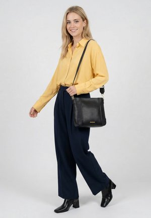 Jeune femme en blouse jaune et pantalon marine portant un sac bandoulière en cuir noir, portant des bottines noires, souriante, debout sur un fond gris.