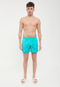 Pantaloni da bagno turchesi con cintura elastica e lacci. Texture liscia, design a lunghezza ginocchio, indossati con sandali bianchi.