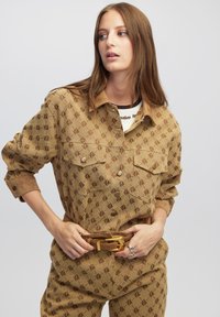 Beige gemusterter Jumpsuit mit Knopfleiste, zwei Brusttaschen und einem braunen Gürtel mit goldenen Beschlägen. Glatte Stofftextur.