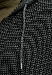 Hoodie noir texturé avec fermeture éclair et cordon de serrage gris avec une extrémité en métal. Le motif en maille gaufrée enrichit la profondeur du tissu.
