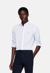Camisa de botones azul claro con un sutil patrón de cuadros y un pequeño logo en el pecho, combinada con pantalones oscuros.