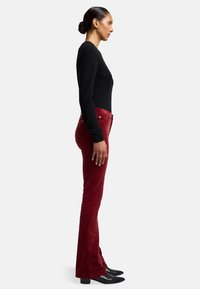 Rode fluwelen broek met uitlopende pijpen en een zachte textuur, gecombineerd met een nauwsluitend zwart longsleeve top en puntige zwarte enkelboots. Profielweergave.