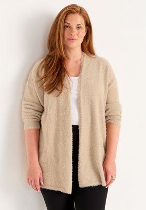 Beige avokardigan med öppen framsida tillverkad av mjukt, texturerat tyg. Har långa ärmar med uppvikta manschetter och en avslappnad passform, stylad över en vit topp.