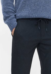 Donkerblauwe broek van een gestructureerde stof, met een trekkoord in de taille en zijzakken. Gecombineerd met een blauw gespikkelde gebreide trui.