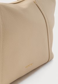 Τσάντα χειρός από μπεζ δερματίνη με υφή, με χρυστό φερμουάρ και μικρό χρυσό λογότυπο "COCCINELLE" στην κάτω δεξιά γωνία.