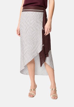 Donna che indossa una gonna bianca asimmetrica con piccoli motivi blu e uno strato marrone con frange, abbinata a sandali beige con tacco e borchie.