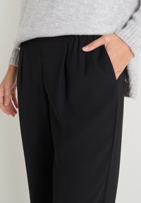 Pantalones negros con una textura suave, cinturilla elástica y bolsillos laterales. Combinados con un suéter de punto acanalado gris claro.