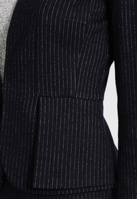 Laivastonsininen liituraitainen bleiseri, jossa istuva leikkaus, lovettu kaulus ja yksi rintatasku. Kangas on sileää ja kudottua.