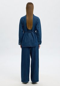 Giacca di denim e pantaloni a gamba ampia in blu scuro. La giacca ha una vita con laccetto, maniche lunghe e una vestibilità comoda. Tessuto liscio e dettagli minimi.