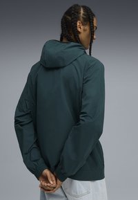 Chaqueta ligera de color verde oscuro con capucha, puños elásticos y un corte relajado. Textura suave con detalles de diseño mínimos.