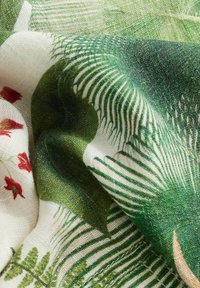 Gros plan sur un tissu avec des motifs botaniques verts et blancs, comprenant des feuilles et de petites fleurs rouges, montrant la texture et les plis.