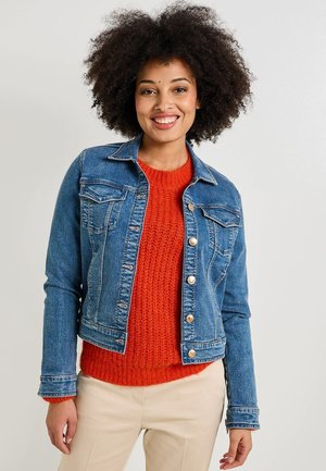 Chaqueta de mezclilla con un diseño ajustado, que presenta dos bolsillos en el pecho, botones de metal y un color azul medio. Lleva puesta una sudadera de punto naranja.