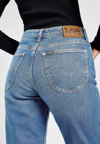 Blauwe denim jeans met een hoge taille, contrasterende gele stiksels en gebogen achterzakken. Heeft een bruine leren merkpatch op de tailleband.